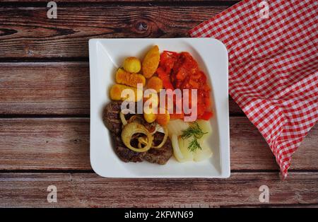 Rindersteak mit Zwiebelringen, Kartoffelkroketten und Gemüse (Fenchel und Paprika) auf einem Teller mit dunklem Holzhintergrund und karierter Serviette. Stockfoto