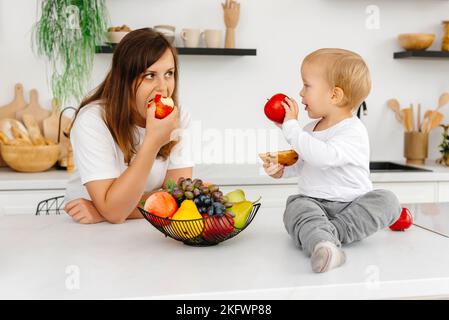 Mom und ihr Sohn sind in der Küche und essen gesundes Essen. Das Baby hält einen Keks und einen Apfel. Reduktierendes Essen Mit Pflanzlicher Nahrung Stockfoto