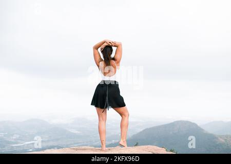 kaukasische reife Frau in weißem Top und schwarzem Rock auf dem Berg stehend mit Händen auf dem Kopf Stockfoto