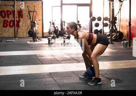 kaukasisches Mädchen, das in einem großen Fitnessstudio Sport treiben und einen schweren Ball mit großer Anstrengung heben kann Stockfoto