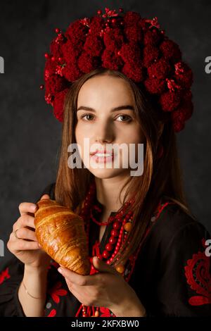Ukrainische Frau in Nationaltracht hält auf dunklem Grund ein Tablett mit rosigen Croissants Stockfoto