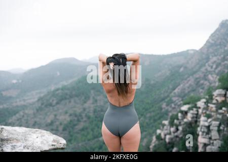 Kaukasische reife Frau mit ihren Händen in ihrem schwarzen Haar von hinten, die die Berglandschaft in einem grauen Badeanzug betrachtet Stockfoto