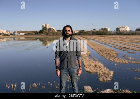 Junger Kaukasier mit langem Bart und grauem Hemd, der mit geschlossenen Augen auf einem von Wasser überfluteten Feld die Kamera ansah Stockfoto