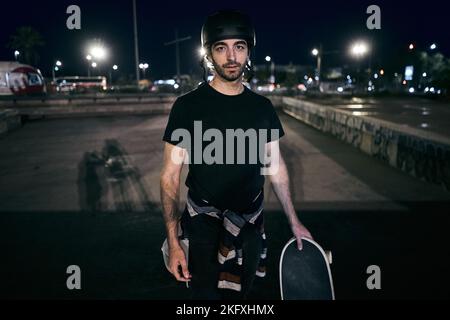 kaukasischer Junge Skater in einem gestreiften Pullover an der Taille gebunden hält ein Skateboard mit seiner Hand in der Stadt Nacht Stockfoto