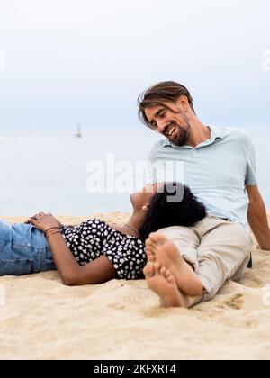 Heterosexuell vielfältiges junges Paar, das liebevoll vor dem Meer sitzt Stockfoto
