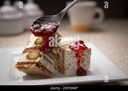 Ein Nahaufnahme-Studiofoto des Löffelens von Erdbeergelee auf Tiramisu-Kuchen auf einem weißen Teller. Stockfoto