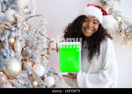 Lächelndes schwarzes Mädchen in Weihnachtsmann Hut zeigt Tablette mit grünem Bildschirm, hält Chroma-Key-Gadget in der Nähe von Weihnachtsbaum, Werbung Weihnachtsartikel, Apps Stockfoto