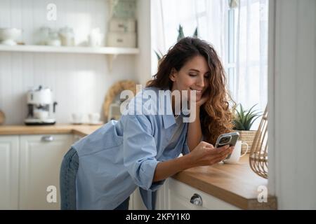 Glückliche, angenehme junge Frau, die mit dem Smartphone in der Küche steht und online nach Kochrezepten sucht Stockfoto