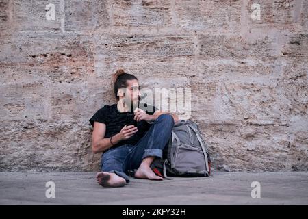 Bärtiger kaukasischer junger Mann barfuß mit schmutzigen Füßen, die auf dem Boden sitzen und sich mit seinem Smartphone in den Händen an eine Steinwand lehnen Stockfoto