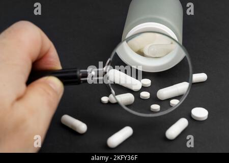 Medizin und Gesundheitskonzept. Auf schwarzem Hintergrund liegen Pillen und ein Glas. Eine Person untersucht sie durch eine Lupe. Stockfoto