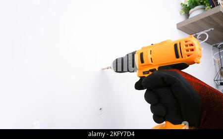 Der Mechaniker verwendet einen elektrischen Schraubendreher oder einen elektrischen Bohrer, um die Mutter an der weißen Wand in einem Haus zu befestigen Stockfoto