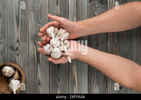 Nahaufnahme gekürzte Hände, die eine Schüssel Knoblauchknollen auf einem Holztisch halten. Gemüsesalat mit würzigen Gewürzen. Kochen einer natürlichen Bio-Farm Stockfoto