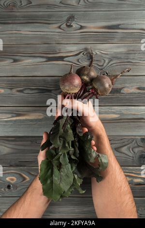 Vertikale, nicht erkennbare, gekürzte Menschenhände, die rohe, frische, saubere Rote Bete mit Stielen und Blättern auf Holzhintergrund halten. Gesunde Ernährung, Diät für Stockfoto