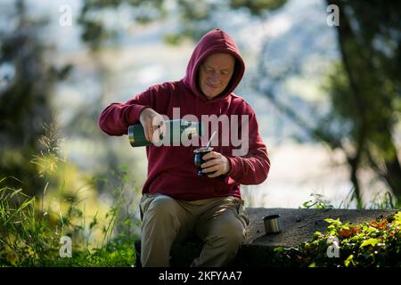 Ein Mann im Park gießt kochendes Wasser aus einer Thermoskanne in einen Mate-Becher. Stockfoto