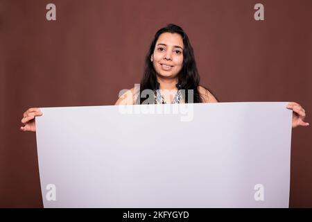 Lächelnde inderin mit leerem Banner mit Platz für die Werbeaktion und Blick auf die Kamera. Modell stehend mit leerem Whiteboard in den Händen, Marketing-Werbung Mockup Stockfoto
