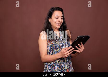 Lächelnde inderin hält digitales Tablet, verwendet Touchscreen und sendet Textnachrichten. Fröhliche Dame, die online spricht, SMS schreibt, lustige Videos auf elektronischem Gadget ansieht, mittellange Aufnahme im Studio Stockfoto