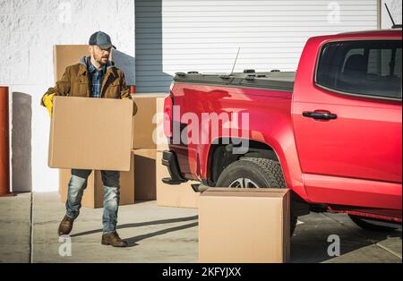 Professioneller Mitarbeiter eines Umzugsunternehmens im mittleren Alter, der eine Kartonschachtel mit den Habseligkeiten des Kunden zu seinem Big Red Pickup Truck trägt. Relocation Services Stockfoto