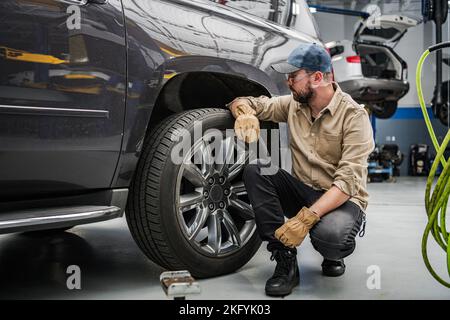 Professioneller Mechaniker mittleren Alters überprüft den Zustand der Autoreifen vor planmäßigen saisonalen Änderungen. Fahrzeugwerkstatt im Hintergrund. Stockfoto