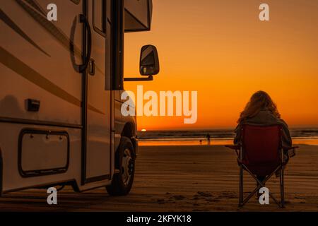 Frau sitzt friedlich auf dem Stuhl am Sandstrand und beobachtet ihren Mann beim Spielen mit Hund im Meer. Moderne Wohnmobil neben geparkt. Landschaftlich Schöner Blick Auf Den Sonnenuntergang. Stockfoto