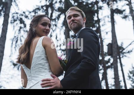 Gerade verheiratet Wald Fotoshooting. Mittleres Porträt einer langhaarigen, brünette türkischen Braut und ihres skandinavischen Mannes, die mit einem Lächeln auf ihren Gesichtern auf die Kamera zurückblicken. . Hochwertige Fotos Stockfoto
