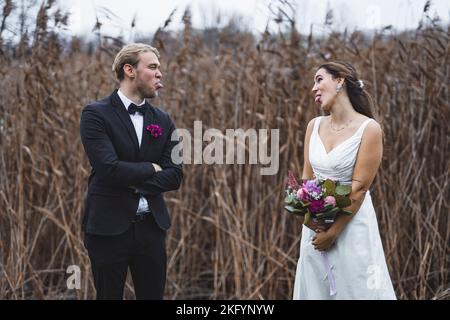 Lustige Hochzeitserinnerungen. Die Braut aus dem Nahen Osten in schönem weißen Kleid und ihr kaukasischer Ehemann in traditionellem Smoking mit Schmetterlingsband, die ihre Zungen aneinander klebten und scherzte. Lachen und Spaß beim Herbstfotoshooting im Freien. Hochwertige Fotos Stockfoto