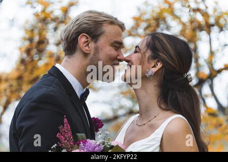 Pure Liebe und Glück des neuvermählten heterosexuellen ethnischen Paares. Die attraktive türkische langhaarige Braut und ihr hübscher skandinavischer Ehemann berühren sich gegenseitig die Nase. Blick nach hinten im Freien. Herbstbäume im Hintergrund. Hochwertige Fotos Stockfoto