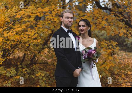 Erstaunliche Farben des Herbstwaldes. Heterosexuelles Paar während der Hochzeit Fotoshooting. Mittellange Aufnahme des kaukasischen Ehegatten und seiner türkischen Frau, die in ehelichen Kleidern nebeneinander stehen. Hochwertige Fotos Stockfoto