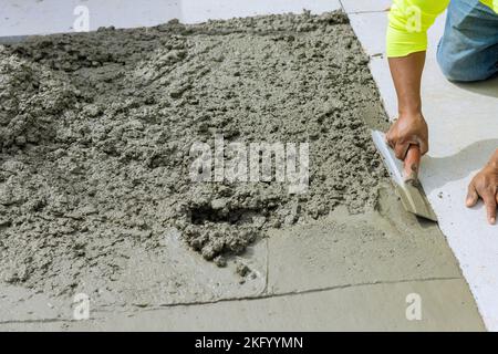 Der Arbeiter hält eine Stahlkelle, während er den neu gegossenen Betonweg im Baubereich glättet und ebnet Stockfoto