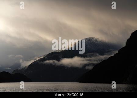 Dramatisches und launisches Morgenlicht beleuchtet einen schneebedeckten Hang im Doubtful Sound. Stockfoto