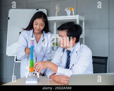 Zwei asiatische Ärzte in weißen Uniformen unterhalten sich und arbeiten im medizinischen Büro zusammen. Der orthopädische Erwachsene männliche Arzt vermittelt Wissen und Beratung Stockfoto