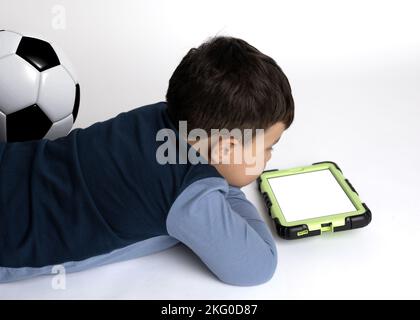 Ein Junge, etwa 4 Jahre alt, und sein Football Ball lag auf dem Boden und schaute sich sein Tablet an. (Leerer Bildschirm) Stockfoto