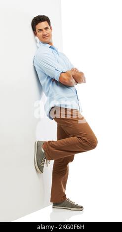 Lässige Mode im Studio. Ein hübscher junger Mann, der sich an einer Wand lehnt, während er auf einem weißen Hintergrund isoliert ist. Stockfoto