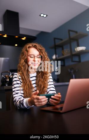 Die ziemlich blonde Frau lenkte sich von der Arbeit auf dem Laptop ab und schaute sich Videos auf dem Smartphone an. Freiberufler mit Mobiltelefon und Surfen, mobil spielen Stockfoto