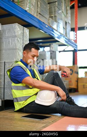 Porträt, müde und verärgert tausendjährigen asiatischen männlichen Lagerarbeiter sitzen im Lager. Industrie-, Ingenieur-, Lager-oder Ladenhausarbeiter. Stockfoto