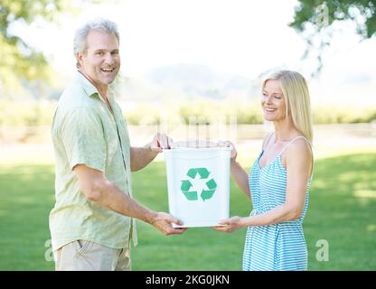 Verantwortung für das übernehmen, was wir zurücklassen. Ein glückliches Paar, das in einem Park steht und einen Recyclingbehälter hält und die Kamera anlächelt. Stockfoto