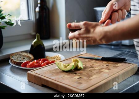 Prozess der Frau schneiden Avocado für Toasts im Kücheninnenraum. Frau bereitet am Morgen ein gesundes Frühstück zu. Vegane Küche Stockfoto