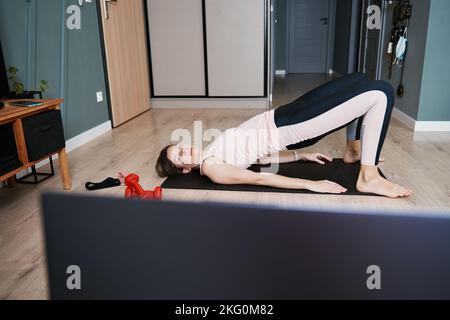 Frau macht Sportübungen im Wohnzimmer. Fernunterricht mit Fitnesstrainer zu Hause, mit Laptop für Videoanruf. Frau in Sportkleidung macht Cardio auf dem Boden Stockfoto