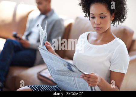 Den Überblick über internationale Entwicklungen behalten. Schöne afrikanische Geschäftsfrau liest eine Zeitung auf einer Couch mit einem Kollegen im Hintergrund. Stockfoto