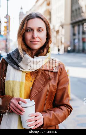 Vertikales Porträt einer jungen blonden Frau, die auf der Straße wartet. Sie trägt eine braune Lederjacke und hält eine wiederverwendbare Tasse Stockfoto