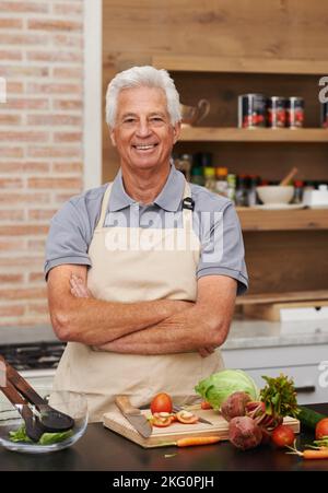 Ich glaube, ich trage diese Küche gut. Ein älterer Mann posiert glücklich in einer Küche, während er eine Schürze trägt. Stockfoto