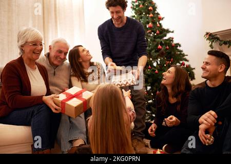 Weihnachten, glücklich und Geschenk mit der Familie im Wohnzimmer für Feiern, Feiertage und festliche Dankbarkeit. Entspannen Sie sich, lächeln Sie und präsentieren Sie sich mit den Eltern, die geben Stockfoto