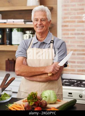 Ich bin jetzt der Meister dieser Küche. Ein älterer Mann, der mit einem Messer in einer Küche posiert. Stockfoto