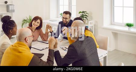 Ein Team glücklicher Geschäftsleute, die am Bürotisch sitzen, die Hände halten und lächeln Stockfoto