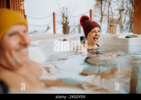 Das ältere Paar mit kitschiger Kappe genießt an kalten Wintertagen gemeinsam die Badewanne im Freien auf der Terrasse. Stockfoto