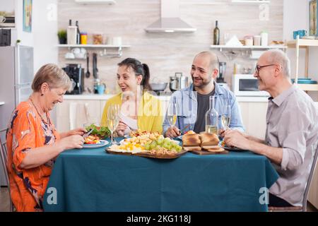 Ältere Frau lachte beim Mittagessen mit der Familie in der Küche. Fröhlicher älterer Vater mit Weinglas. Leckere Kartoffeln. Stockfoto