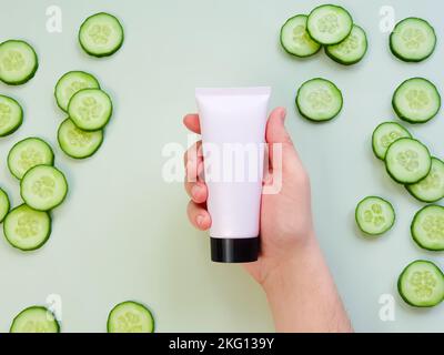 Männliche Hand hält Kunststoff unbranded rosa cremefarbenen Rohr mit schwarzer Kappe. Flasche für Branding und Etikett und frisch geschnittene Gurke auf grünem Hintergrund. Natürlich Stockfoto