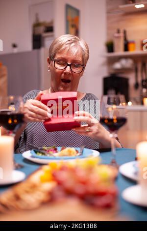 Ältere Frau, die während des festlichen Abendessens schockiert auf Geschenkbox schaut. Fröhliche fröhliche ältere Paar zusammen zu Hause essen, genießen das Essen, feiern ihre Ehe, Überraschungsurlaub Stockfoto