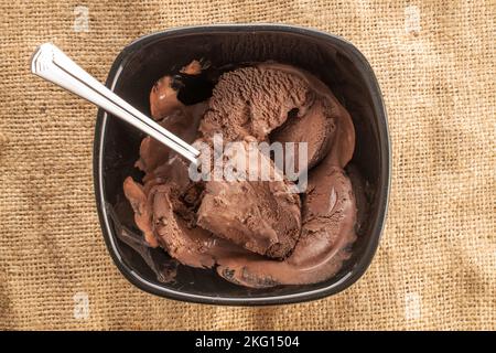 Dunkelbraunes Schokoladeneis in einer schwarzen Keramikschale mit einem Metalllöffel auf einer Jute-Serviette, Nahaufnahme, Draufsicht. Stockfoto