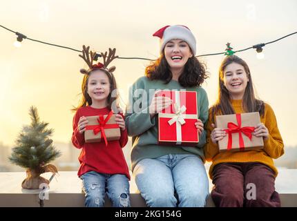 Frohe Weihnachten und frohe Feiertage. Süße kleine Kinder, deren Mutter einen Weihnachtsmann-Hut trägt, präsentieren Geschenke im Freien. Die Familie genießt den Urlaub Stockfoto