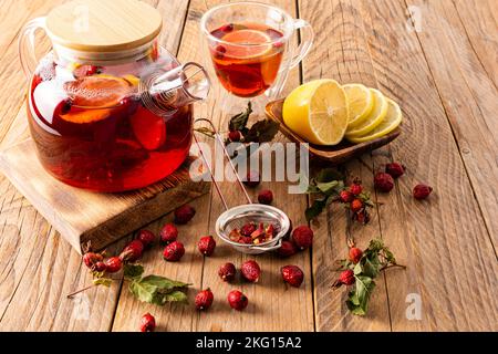 Eine große Glas-Teekane mit Hagebutte und Zitrone auf einem hölzernen Dorftisch mit trockenen Hagebutten. Gesundes Getränk Stockfoto